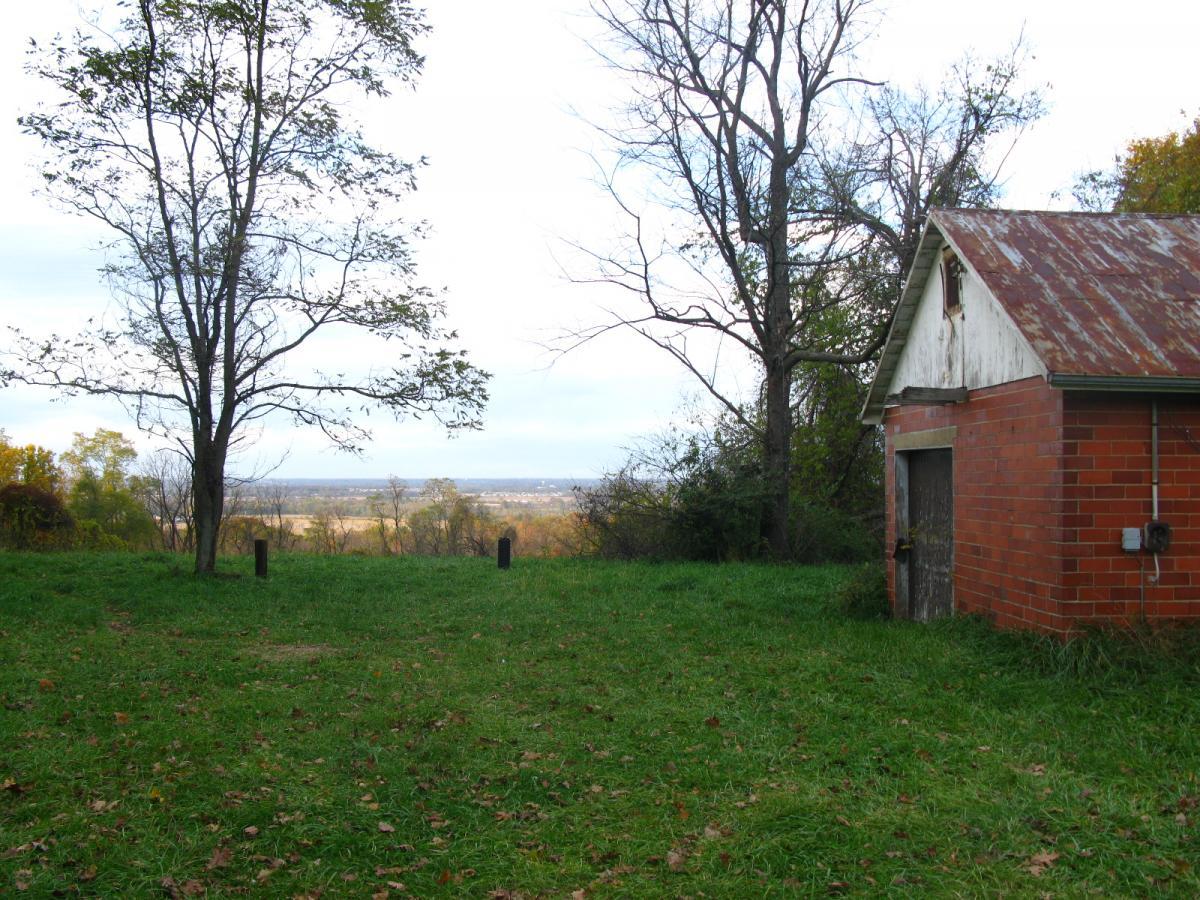 A scenic view of a grassy area with an old brick shed on the right, surrounded by trees. In the background, a landscape with distant hills and a cloudy sky is visible. Chestnut Ridge mountain bike trail.