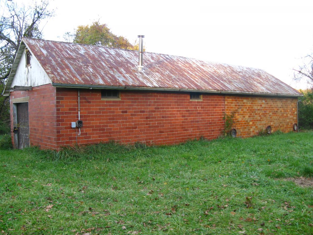 A rustic brick building with a sloped, rusty metal roof, situated in a grassy area surrounded by trees. The exterior features a wooden door and a vent pipe on the roof. The building shows signs of age with some overgrown vegetation nearby. Chestnut Ridge mountain bike trail.