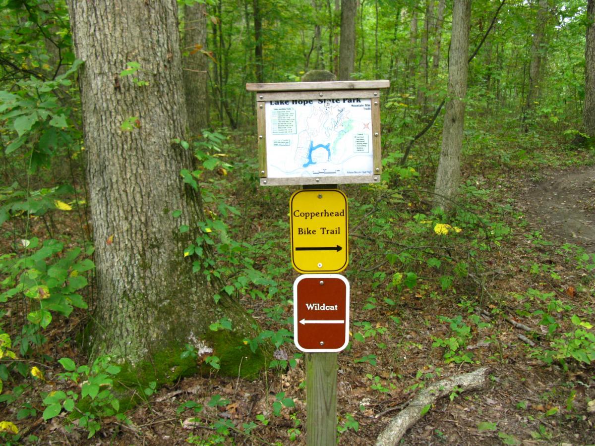 Signpost at Lake Hope State Park displaying a trail map and directions for the Copperhead Bike Trail and Wildcat trail, surrounded by a lush forest with green foliage. Lake Hope State Park mountain bike trail.