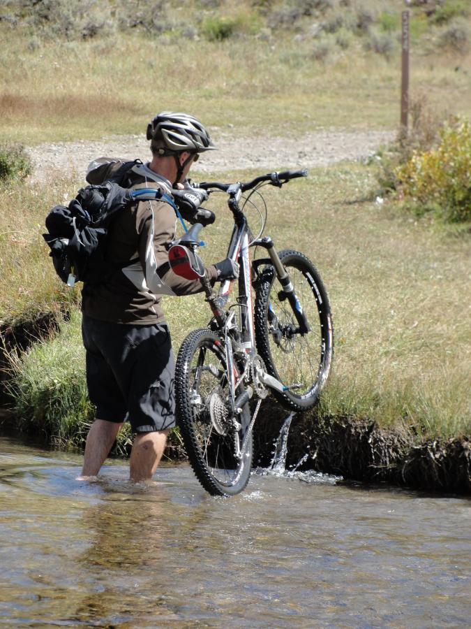 A person wearing a cycling helmet and backpack is walking through a shallow stream while carrying a mountain bike. The surrounding area features grassy fields and shrubs, with a dirt path visible in the background. Doctor Park mountain bike trail.