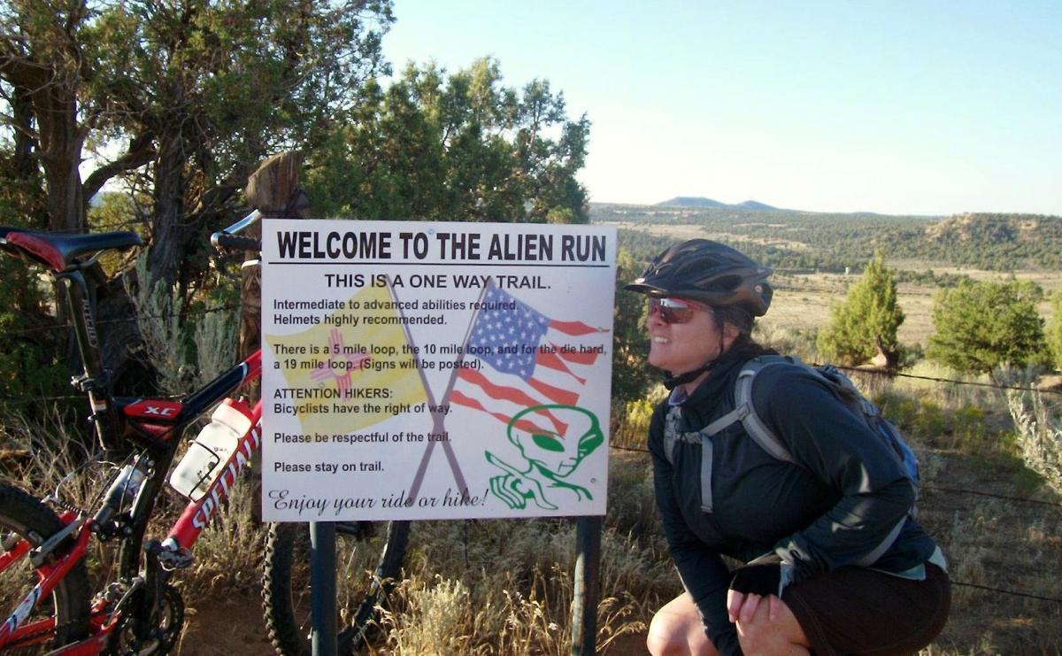 A woman in biking gear smiles near a trail sign that reads "Welcome to the Alien Run." The sign includes information about the trail being one-way, with varying loop lengths and a note for hikers about sharing the trail with cyclists. In the background, there are trees and a scenic landscape. Alien Run Trail mountain bike trail.