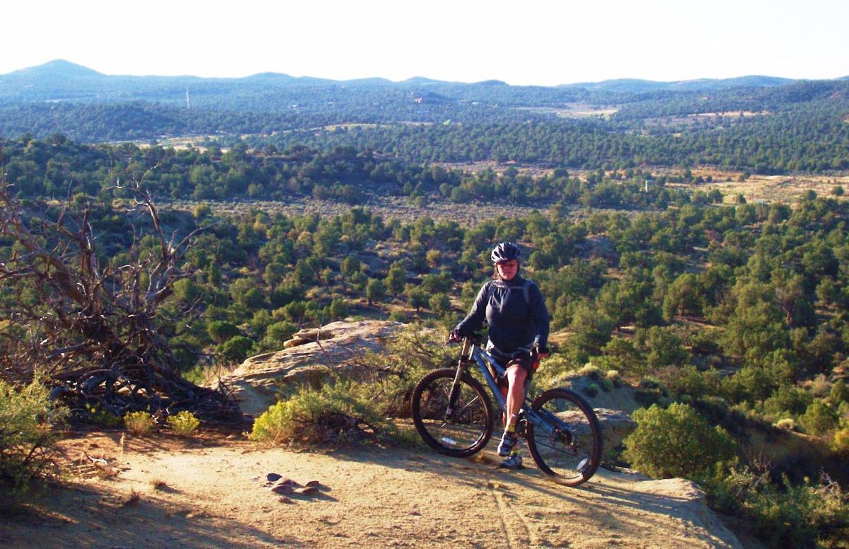 A person in a bike helmet and a dark sweater stands next to a mountain bike on a rocky outcrop, surrounded by a vast landscape of rolling hills and trees. The sun casts a warm glow over the scene, highlighting the rugged terrain and distant mountains in the background. Alien Run Trail mountain bike trail.