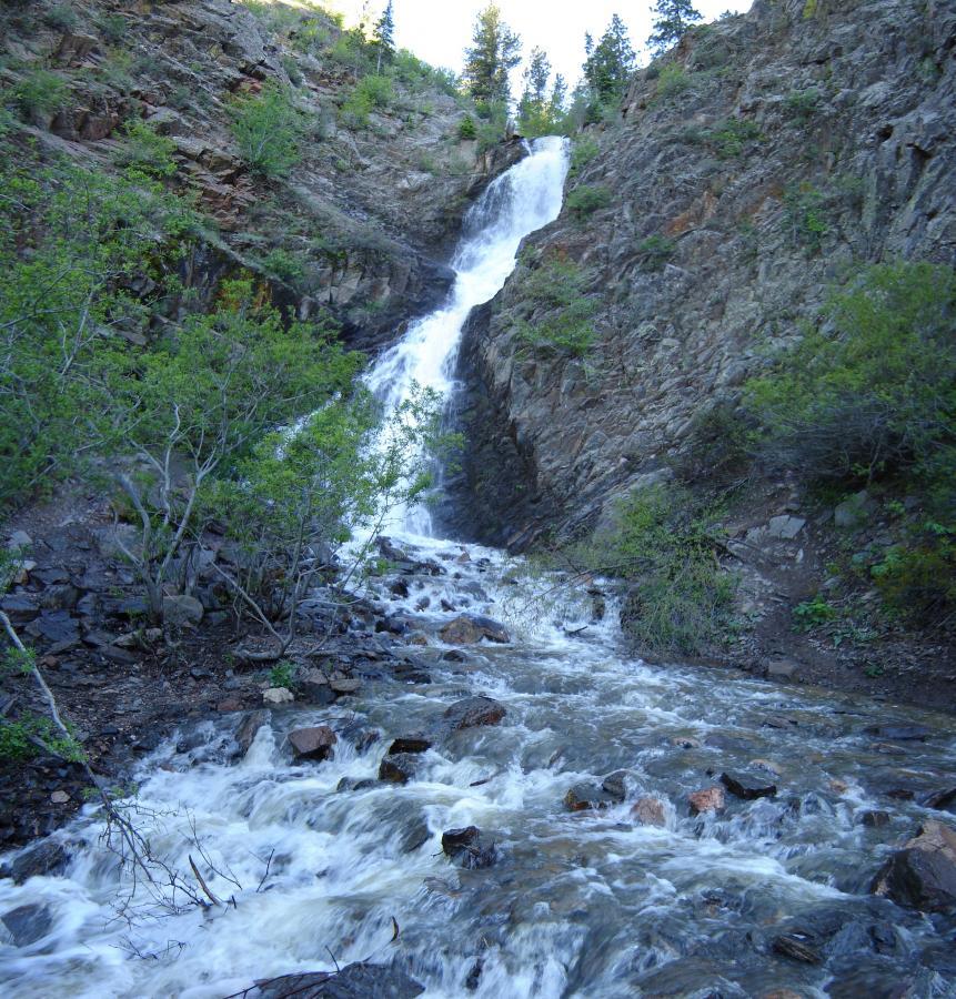 A cascading waterfall flowing down a rocky hillside, surrounded by lush greenery and trees. The water creates a foamy rush as it tumbles over the rocks, with sunlight illuminating the natural landscape. Bridle Trail mountain bike trail.