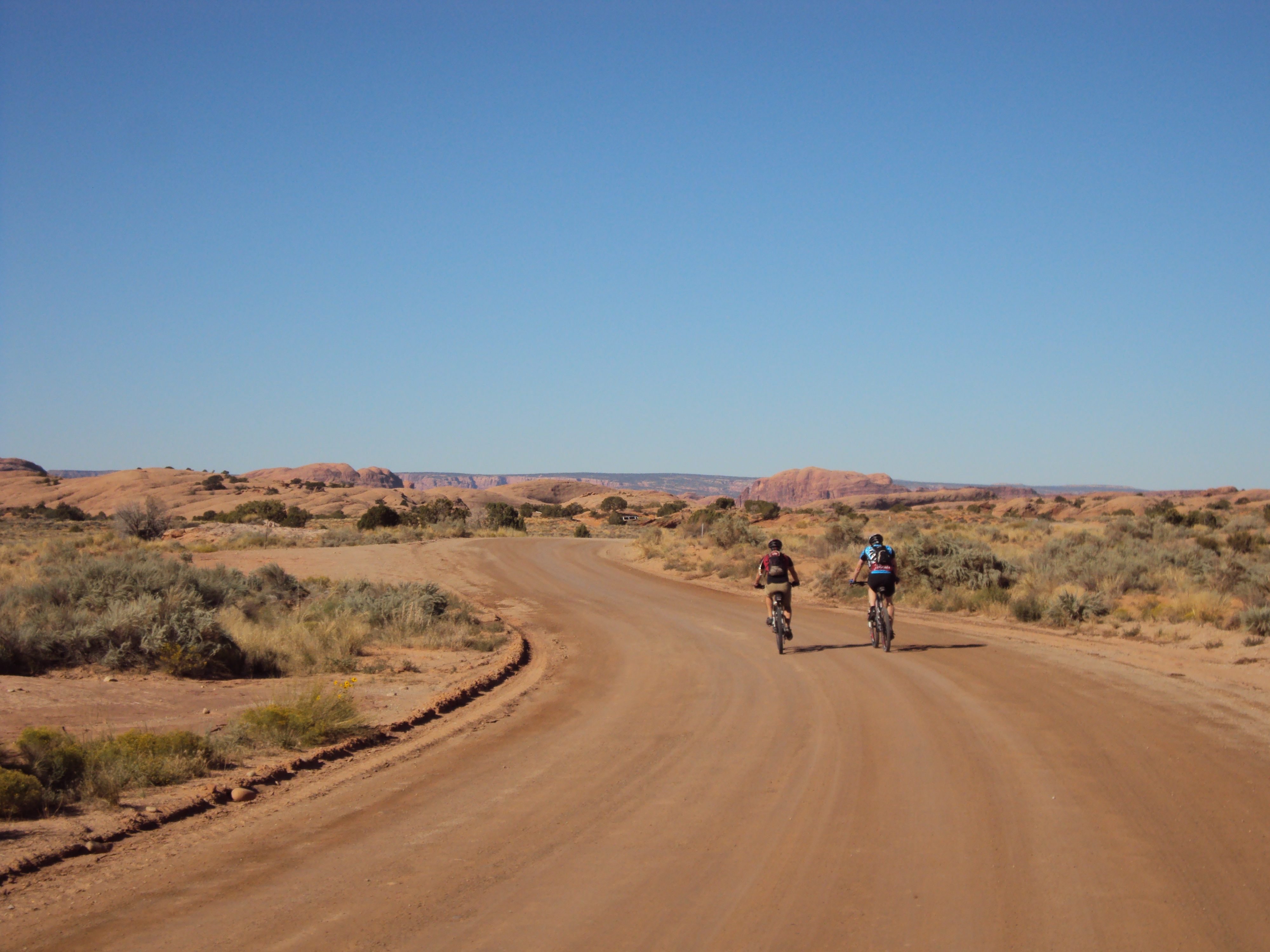 Two cyclists riding along a winding dirt road in a desert landscape, with sparse vegetation and red rock formations in the background under a clear blue sky.