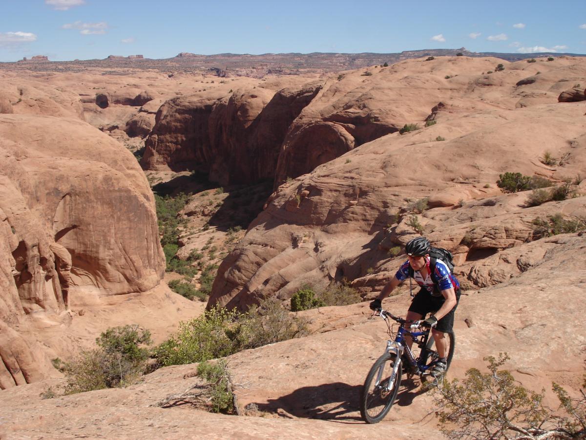 A mountain biker navigates rocky terrain in a desert landscape, surrounded by canyons and distant rock formations under a clear blue sky. Slickrock mountain bike trail.