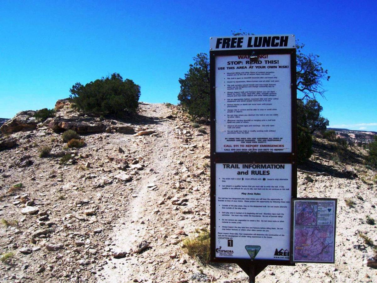 A sign labeled "FREE LUNCH" stands at the beginning of a rocky trail. The sign features important warnings and rules for visitors, advising them to use the area at their own risk. It includes emergency contact information and trail guidelines. In the background, the landscape shows a clear blue sky and shrubbery along the rocky path. Lunch Loops mountain bike trail.