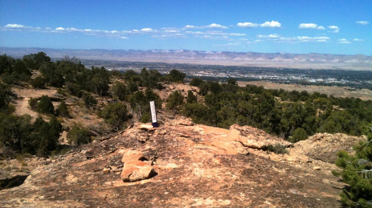 A panoramic view from a rocky hillside, showing a landscape with distant mountains and a valley below. In the foreground, there is a rock surface and a sign that reads "DROP." The area is dotted with shrubs and trees under a clear blue sky with a few clouds scattered. Lunch Loops mountain bike trail.