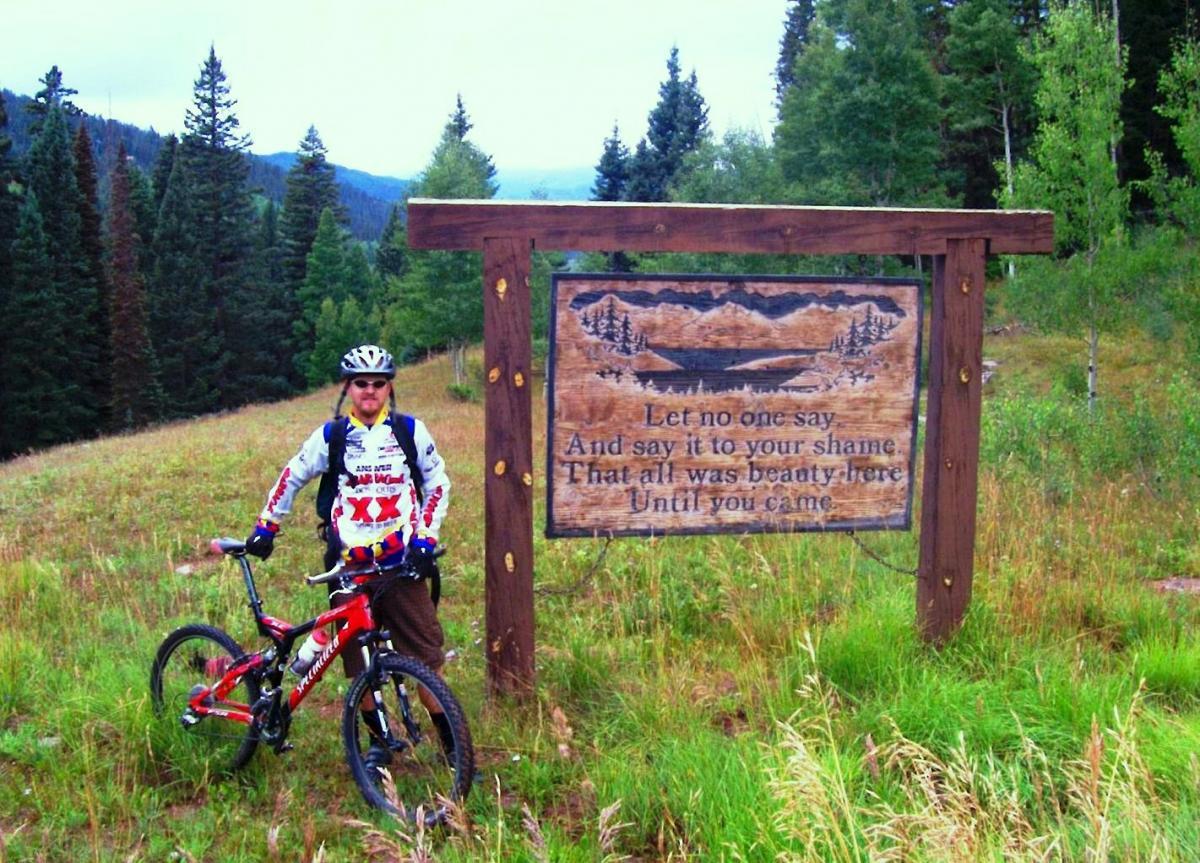 A mountain biker stands next to a wooden sign surrounded by greenery and trees. The sign features an engraved message about beauty, set against a mountainous backdrop. The biker is wearing a helmet, sunglasses, and colorful cycling gear, with a red mountain bike beside him. Hermosa Creek Trail mountain bike trail.