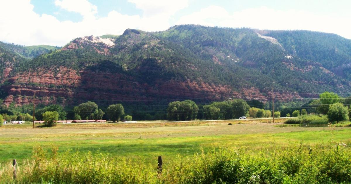 A scenic view of a mountainous landscape featuring reddish rock formations against green hills, with a foreground of grassy fields and scattered trees. The sky is partially cloudy, and a road with vehicles is visible at the base of the mountains. Hermosa Creek Trail mountain bike trail.