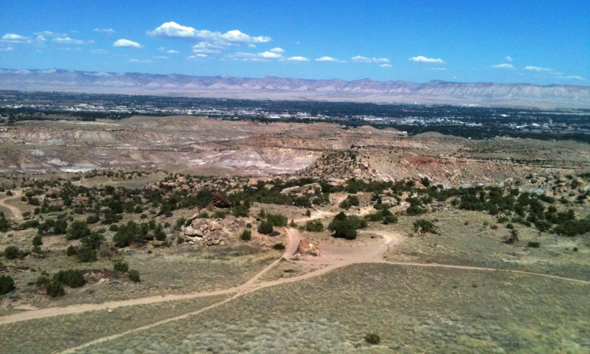 A panoramic view of a rugged landscape featuring rolling hills, sparse vegetation, and distant mountains under a clear blue sky with a few clouds. A dirt path winds through the foreground, leading towards the horizon where a small town is visible below. Lunch Loops mountain bike trail.