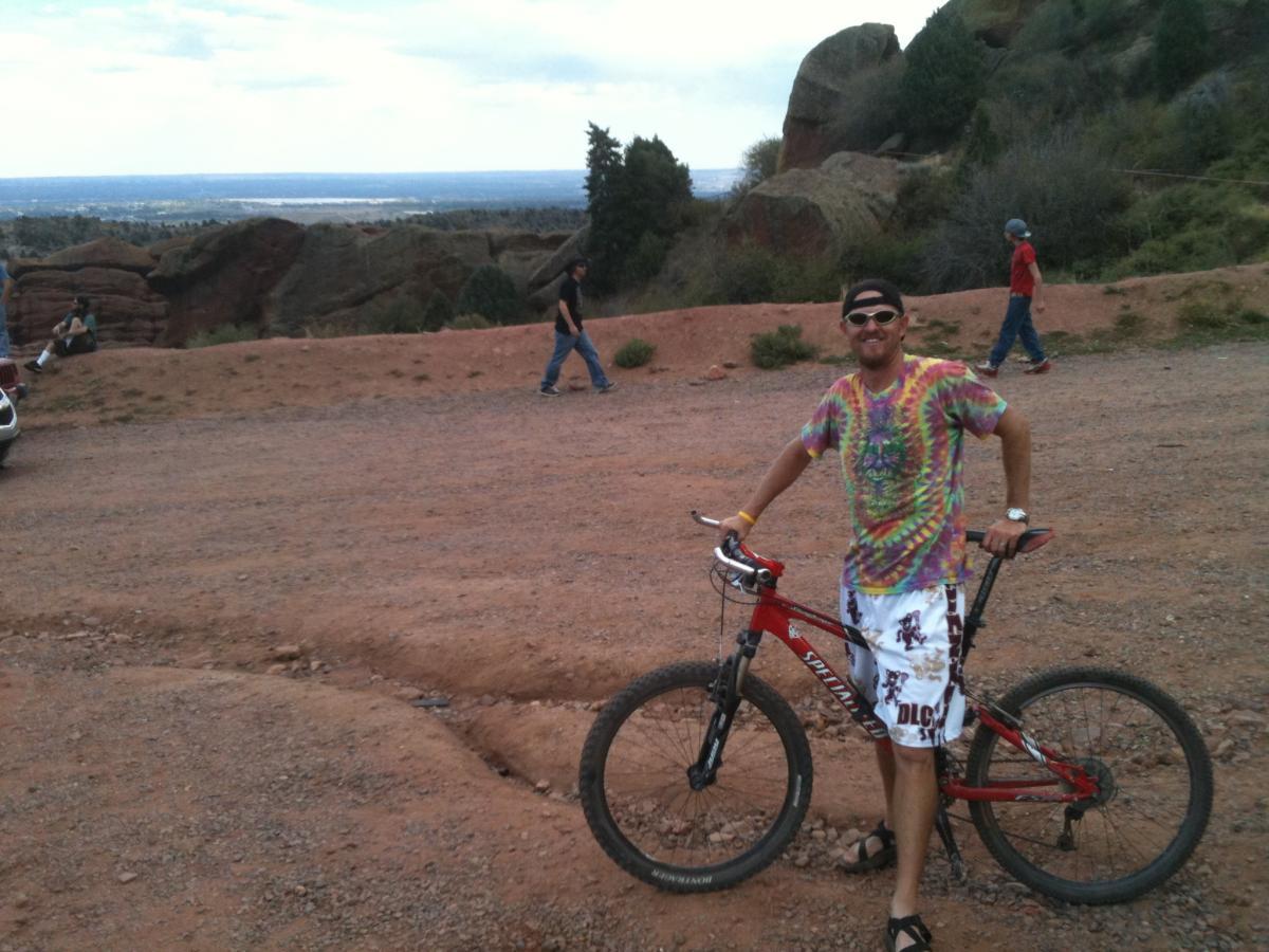 A man wearing a colorful tie-dye shirt and shorts stands beside a red mountain bike on a gravel trail with a view of a valley in the background. Several people can be seen walking in the distance, surrounded by rocky terrain and greenery. Red Rocks / Dakota Ridge mountain bike trail.