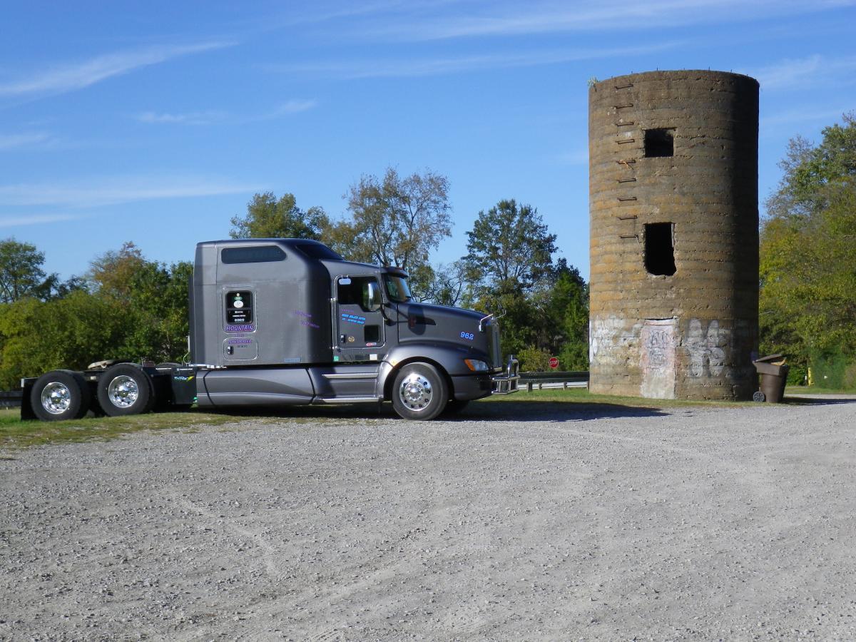 A silver semi-truck parked on gravel next to an old, cylindrical stone tower with two windows, surrounded by greenery and a clear blue sky. There are some trees in the background and a small garbage bin beside the tower. Lock 4 mountain bike trail.