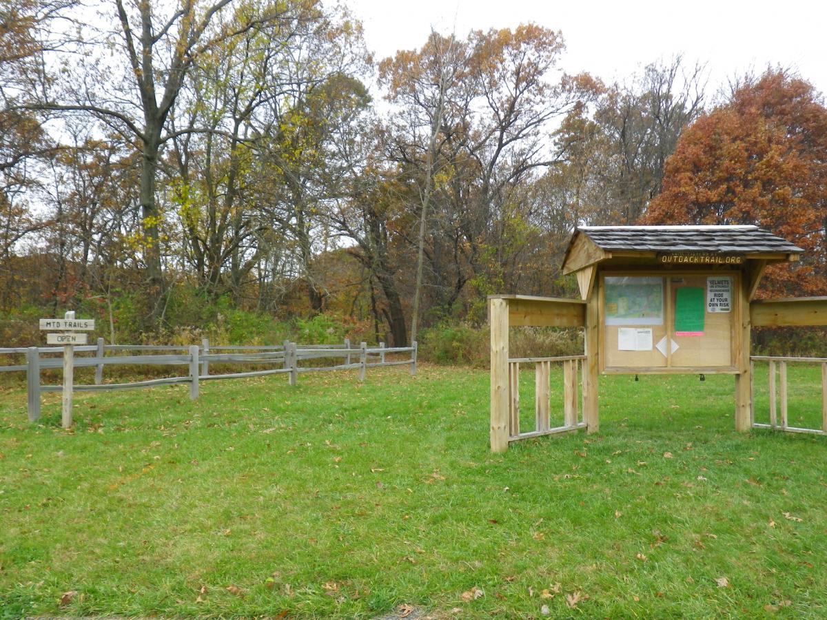 A wooden information kiosk stands in a grassy area, surrounded by trees with autumn foliage. To the left, a sign indicating "MTB TRAILS OPEN" points the way to marked trails. The kiosk has a roof and displays a map along with notices, inviting visitors to explore the outdoor trails. The scene captures a serene outdoor setting during fall. Outback Trail at Imagination Glenn mountain bike trail.