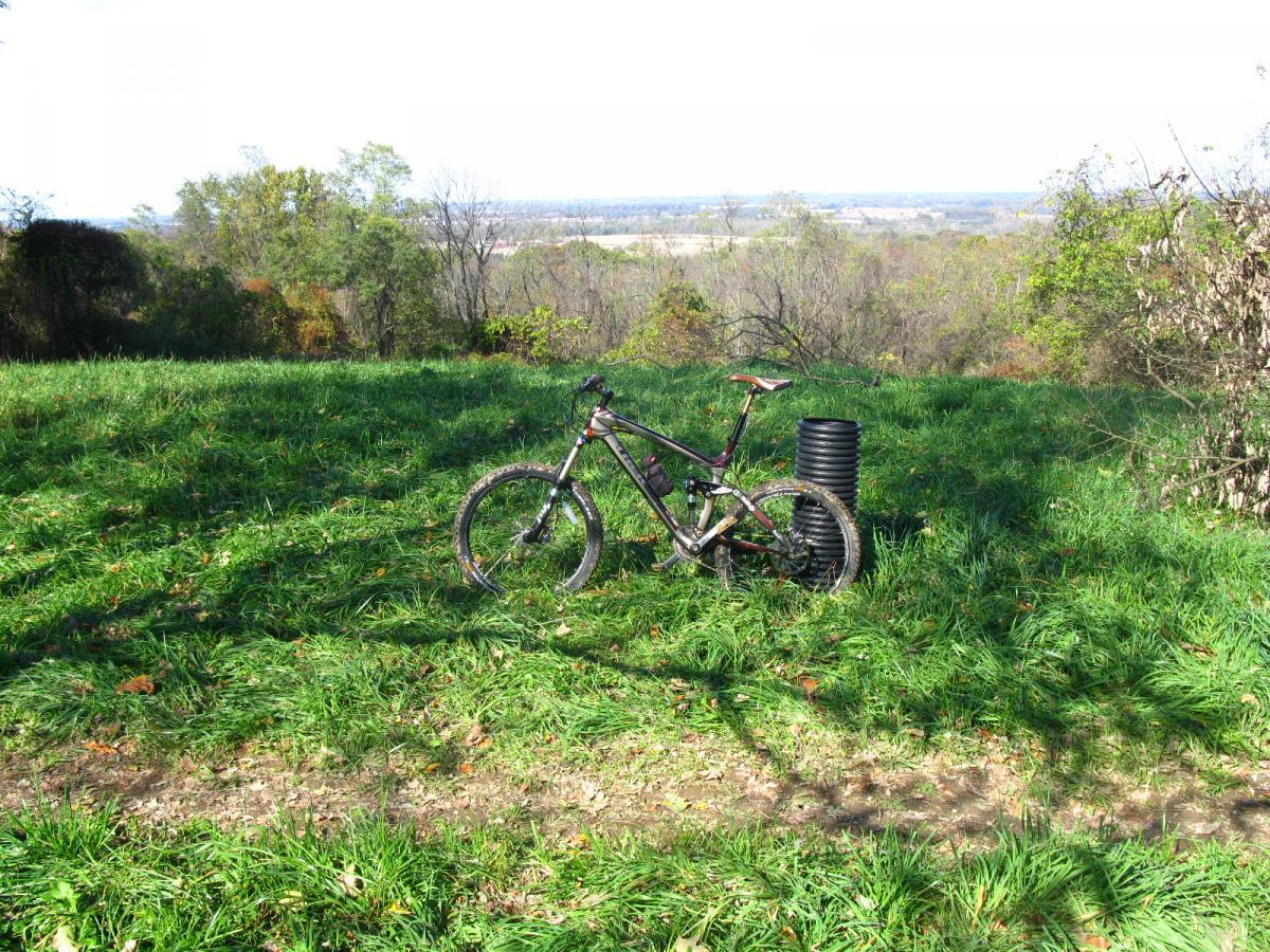 A mountain bike resting on a grassy area with a distant view of rolling hills and trees. A black cylindrical object is positioned nearby. The scene is illuminated by bright natural light, suggesting a clear day. Chestnut Ridge mountain bike trail.