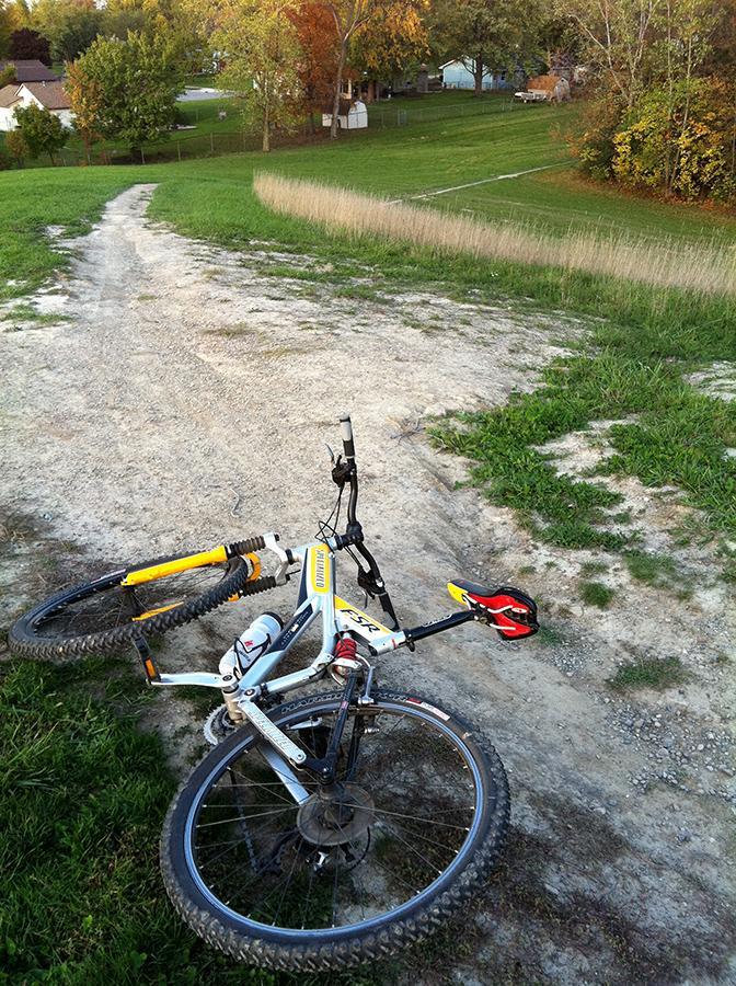 A mountain bike lying on its side on a dirt path, with green grass and trees in the background. The scene captures a winding trail leading through a grassy area into the distance. Franke Park mountain bike trail.