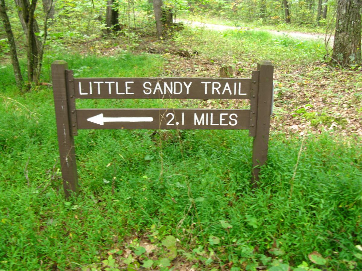 Wooden sign for Little Sandy Trail indicating a distance of 2.1 miles, with an arrow pointing left, surrounded by greenery in a forested area. Lake Hope State Park mountain bike trail.