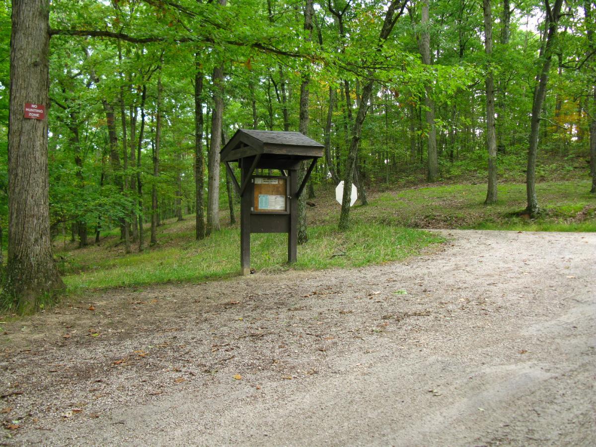 A wooden information kiosk situated along a gravel path in a green forested area, surrounded by trees. A signpost labeled "No Hunting Zone" is visible on a nearby tree, with greenery and a gentle slope in the background. Lake Hope State Park mountain bike trail.