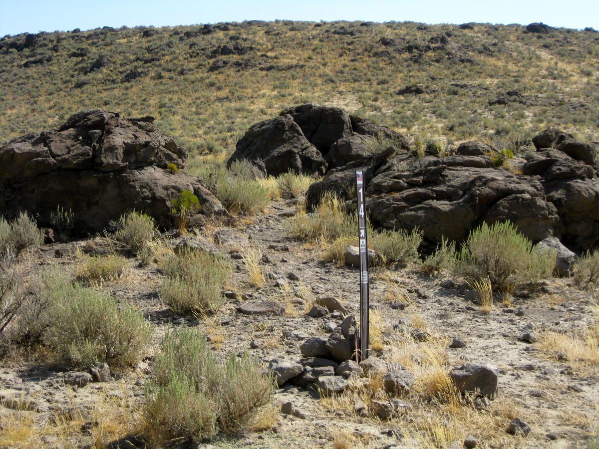 A rocky landscape featuring large boulders and sparse vegetation, with a wooden post marked "14" on the right side. The scene is set against a gently sloping hill in the background, under a clear blue sky. Copper Basin Trail 14 & 15: The Playground mountain bike trail.