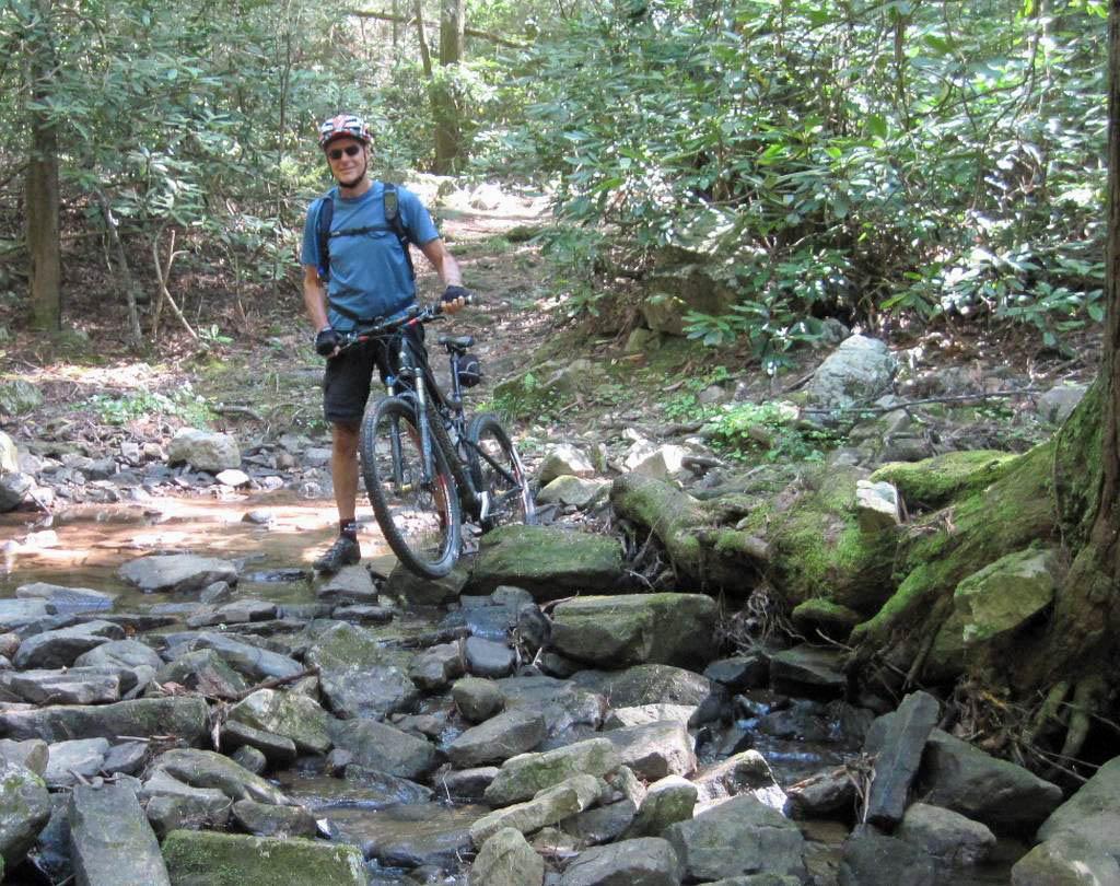 A man stands on rocky terrain beside a shallow stream, holding a mountain bike. He is wearing a blue T-shirt, shorts, and a helmet, surrounded by lush greenery in a wooded area. Sunlight filters through the trees, creating a serene outdoor scene. Rimrock mountain bike trail.