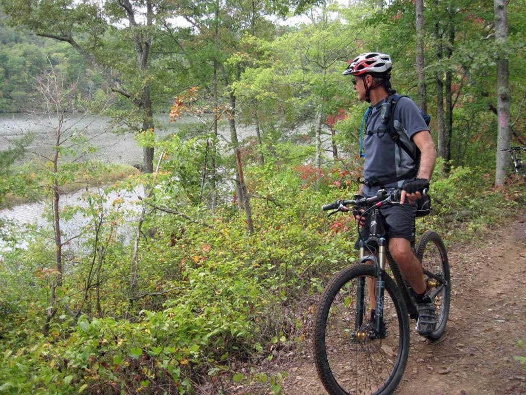 A mountain biker standing next to a bike on a dirt trail, overlooking a lake surrounded by greenery. The scene features trees and shrubs in various shades of green and hints of autumn colors, suggesting a peaceful outdoor setting. Brush Creek mountain bike trail.
