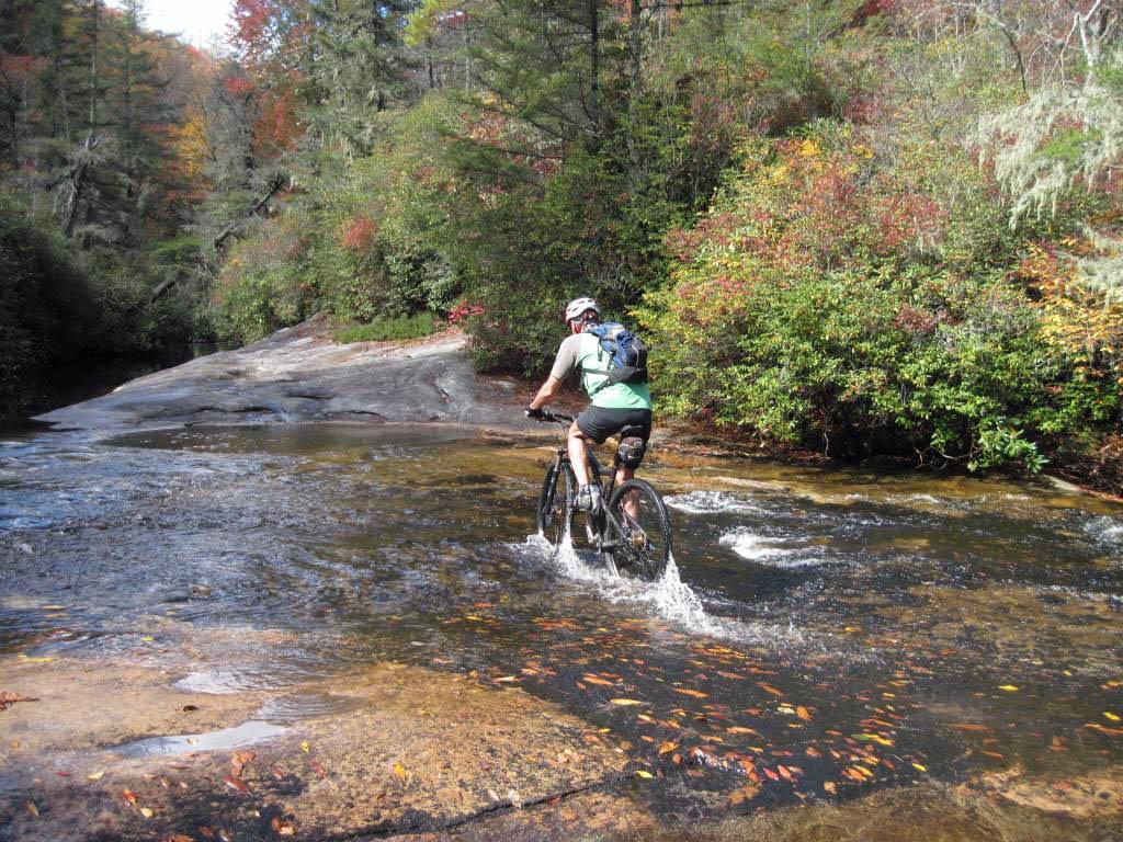 A person riding a mountain bike through a shallow stream, surrounded by vibrant fall foliage and rocky terrain. The cyclist is wearing a helmet and a backpack, with splashes of water visible as the bike navigates the stream. Panthertown Valley mountain bike trail.