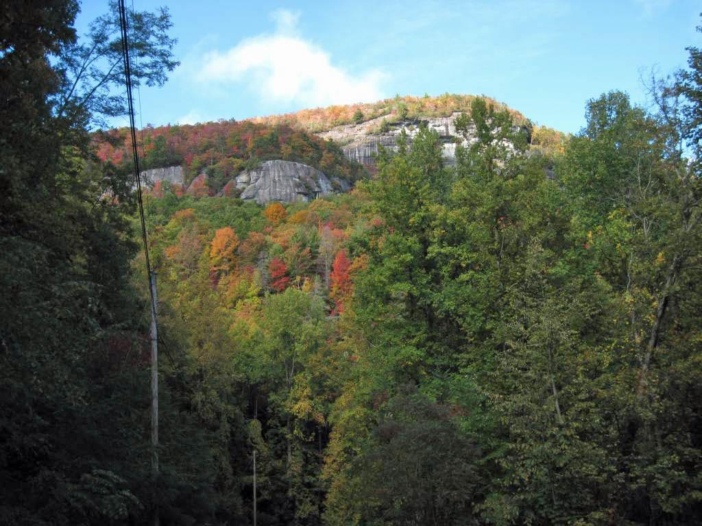 Scenic view of a mountain with a rocky summit, surrounded by vibrant autumn foliage in various shades of red, orange, and green under a clear blue sky. Panthertown Valley mountain bike trail.