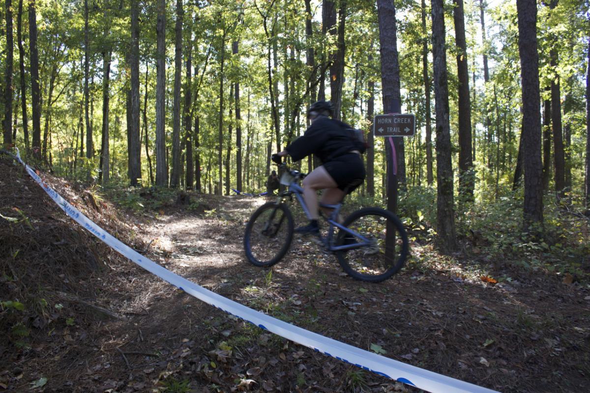 A mountain biker riding along a dirt trail in a forest, with a sign indicating "Horn Creek Trail" visible nearby. The bike is in motion, and the scene is surrounded by tall trees and foliage, suggesting an outdoor recreational setting. Lick Fork (Horn Creek) mountain bike trail.