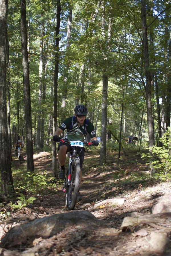 A cyclist navigating a rocky trail in a wooded area, wearing a helmet and cycling gear, while other cyclists can be seen in the background. The trail is surrounded by trees and foliage, indicating a natural outdoor setting. Lick Fork (Horn Creek) mountain bike trail.