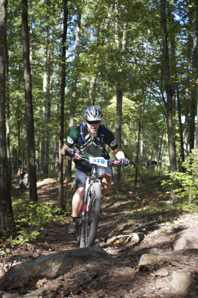 A mountain biker navigating a rocky trail in a forested area, wearing a helmet and cycling gear, with a race number visible on the front of their bike. Sunlight filters through the trees, highlighting the natural scenery.