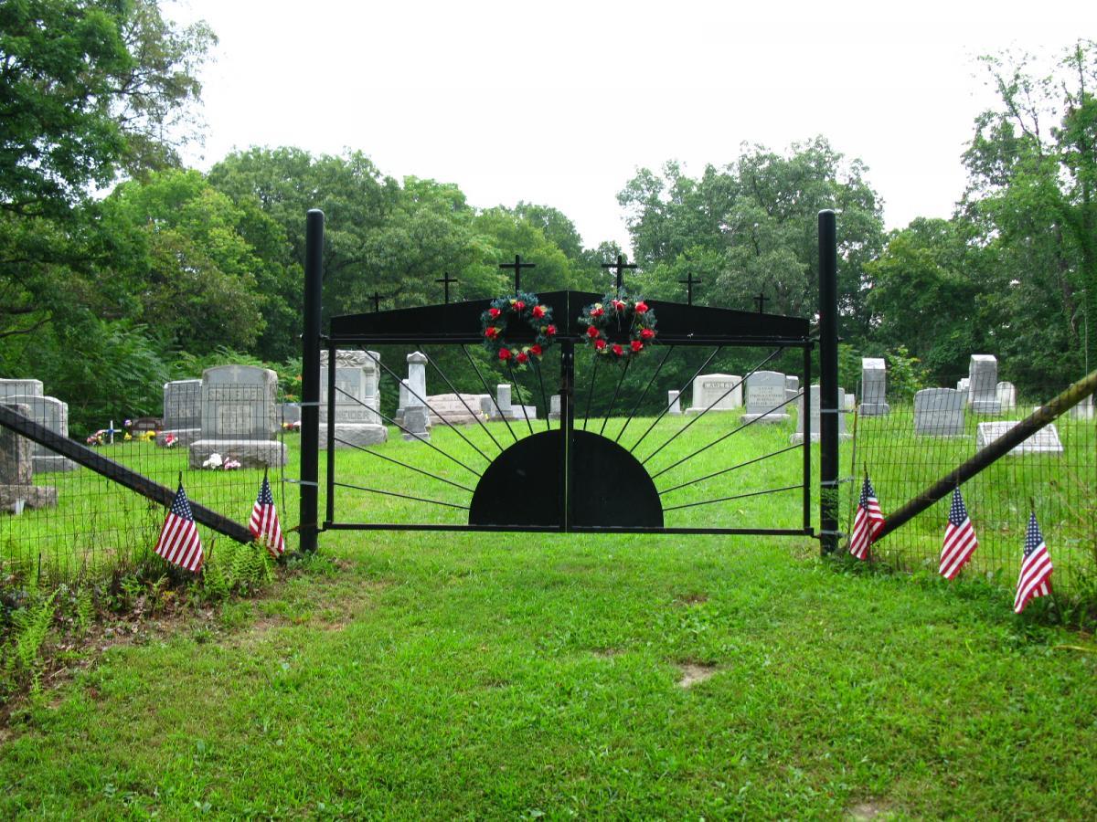 A wrought iron gate leading into a cemetery, adorned with decorative wreaths and flanked by small American flags. The background features several gravestones surrounded by green grass and trees, under a cloudy sky. Archers Fork Loop Trail mountain bike trail.