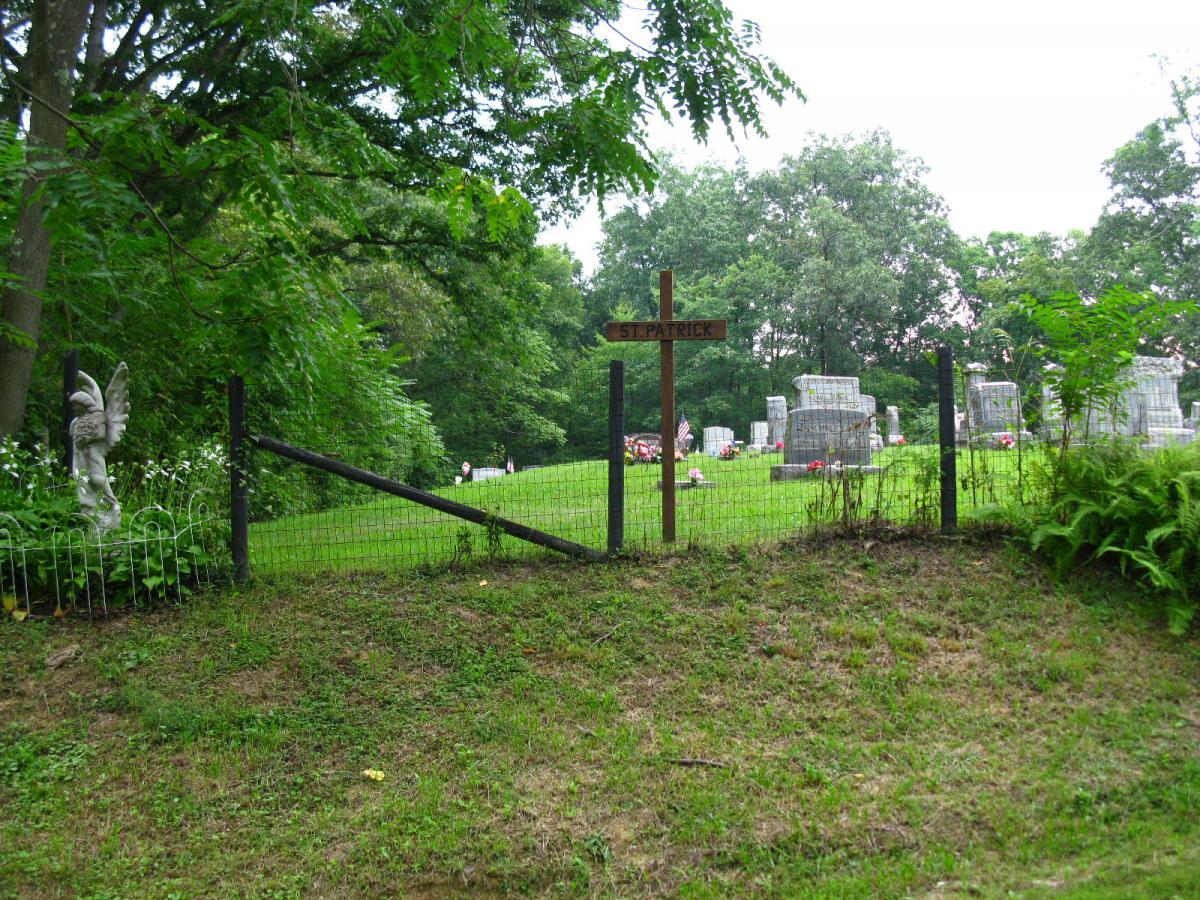 A grassy area leading to a cemetery, marked by a wooden sign that reads "St. Patrick." A fence with a gate frames the entrance, while green trees and bushes surround the site, adding a serene natural backdrop. In the distance, gravestones are visible, some adorned with flowers and small American flags. Archers Fork Loop Trail mountain bike trail.