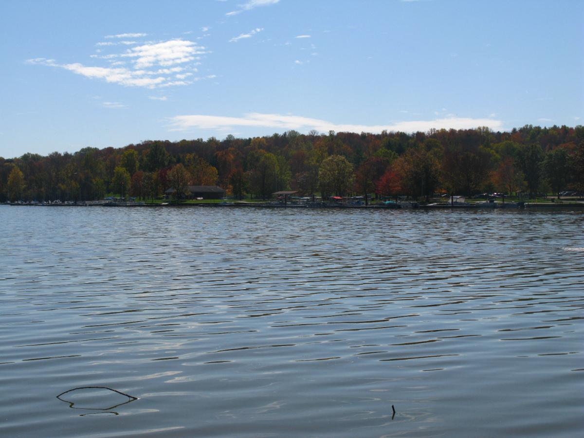 A tranquil lake view with gentle waves, surrounded by trees displaying autumn foliage in vibrant colors. A wooden structure is visible along the shoreline, with a small marina in the background. The sky is clear with a few scattered clouds, creating a serene and scenic atmosphere. Hueston Woods State Park mountain bike trail.