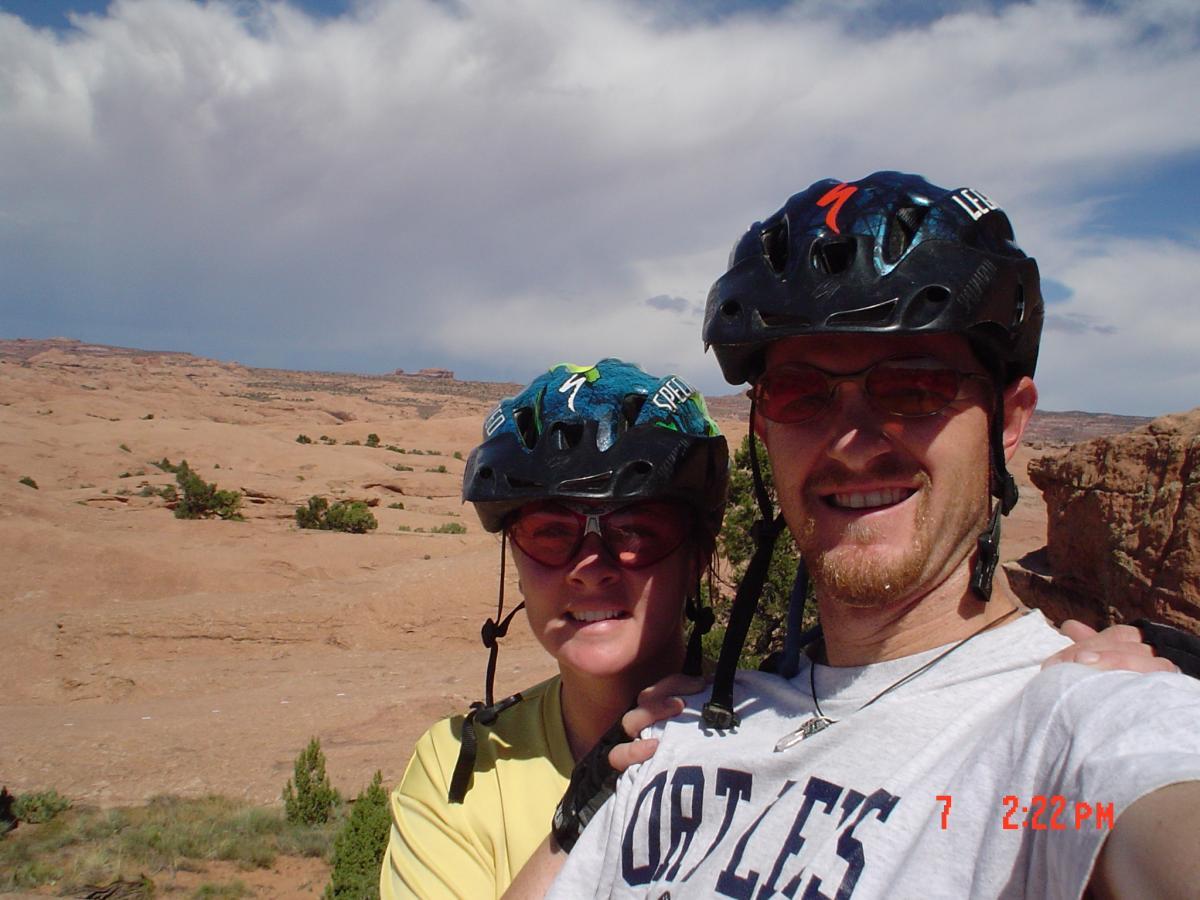 A couple takes a selfie while wearing helmets and sunglasses, against a backdrop of desert landscape with rocky formations and blue sky. Slickrock mountain bike trail.
