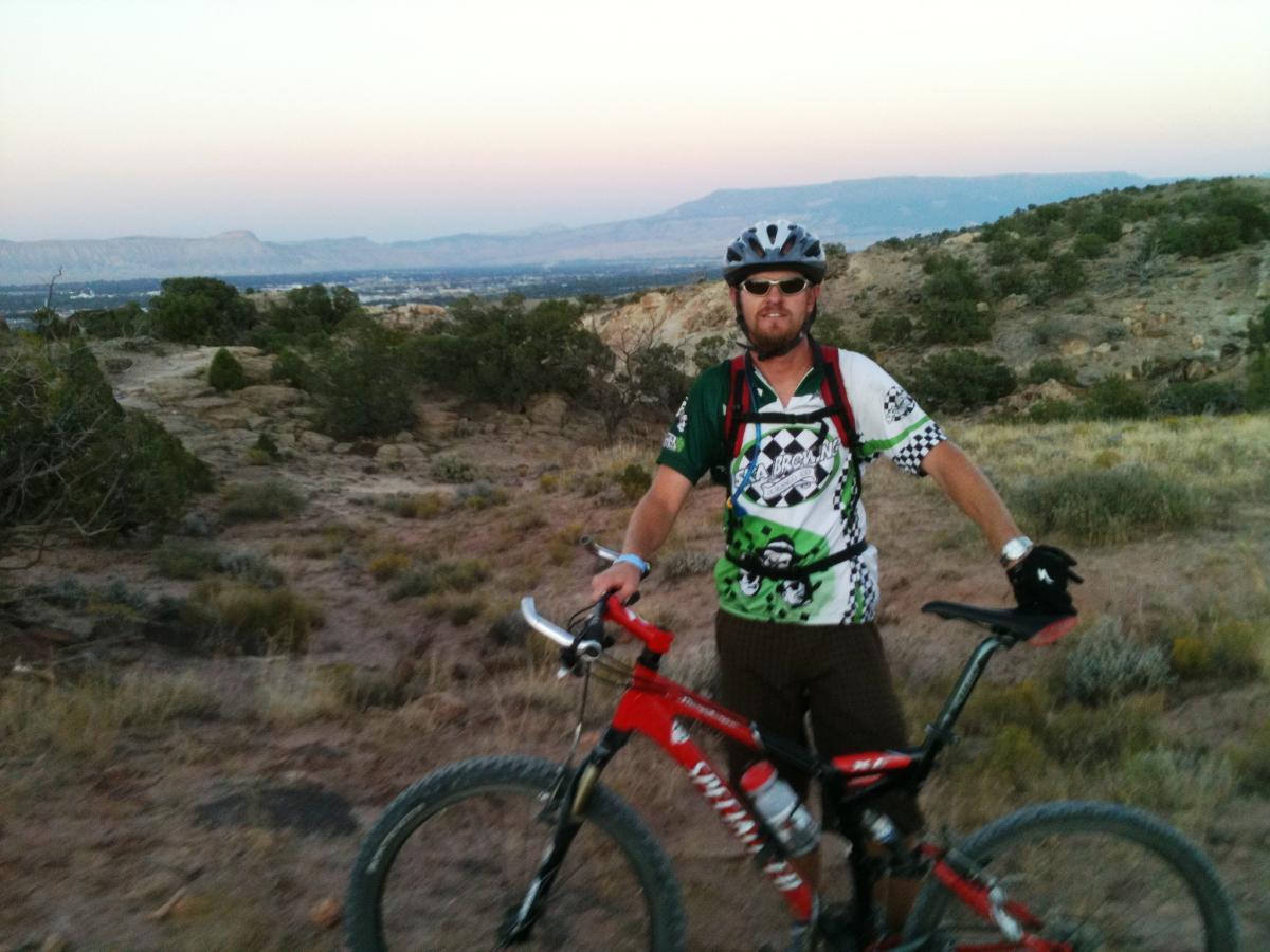 A mountain biker stands next to a red bike on a rugged landscape at dusk. He is wearing a colorful cycling jersey and a helmet, with a varied terrain of shrubs and rocky outcrops in the background, depicting a scenic and adventurous outdoor setting. Lunch Loops mountain bike trail.