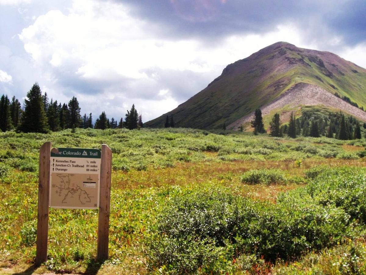A scenic view of the Colorado Trail with a wooden trail sign displaying directions and distances to nearby landmarks. In the background, a prominent green mountain rises under a partly cloudy sky, surrounded by lush vegetation and trees. Colorado Trail: Kennebec Pass To Junction Creek mountain bike trail.
