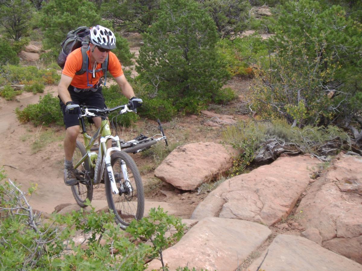 A mountain biker wearing a helmet, orange shirt, and backpack navigates a rocky trail surrounded by greenery and trees. The Whole Enchilada mountain bike trail.