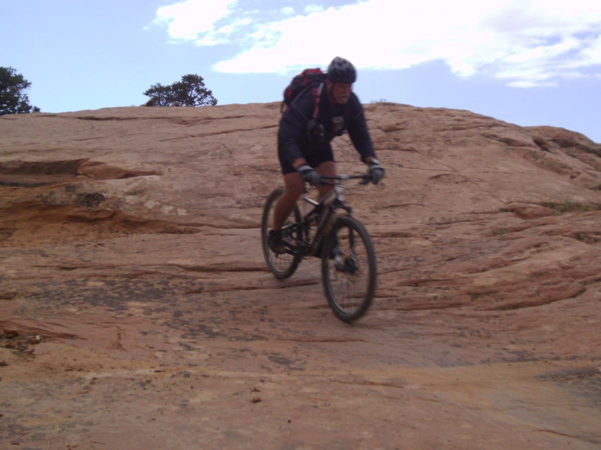 A mountain biker riding down a rocky slope with a rugged terrain, surrounded by a blue sky and sparse trees in the background. The Whole Enchilada mountain bike trail.
