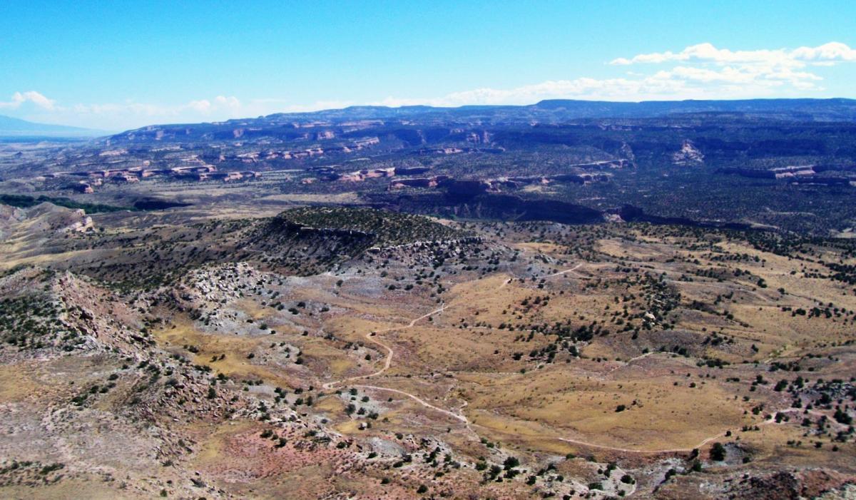A panoramic view of a rugged, arid landscape featuring rolling hills, scattered shrubs, and distant mesas under a clear blue sky. Moore Fun mountain bike trail.
