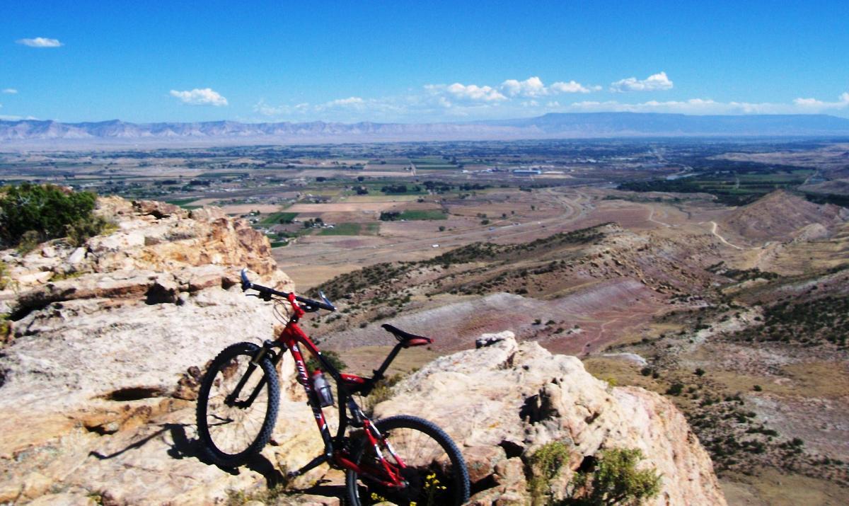 A mountain bike leaning against a rocky outcrop, overlooking a vast landscape of rolling hills and fields under a clear blue sky with scattered clouds. Moore Fun mountain bike trail.