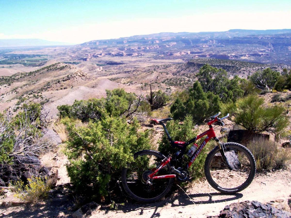 A mountain bike rests next to a rocky outcrop, surrounded by sparse vegetation and a vast mountainous landscape in the background. The sky is bright and clear, showcasing the expansive view of rolling hills and distant cliffs under sunlight. Moore Fun mountain bike trail.