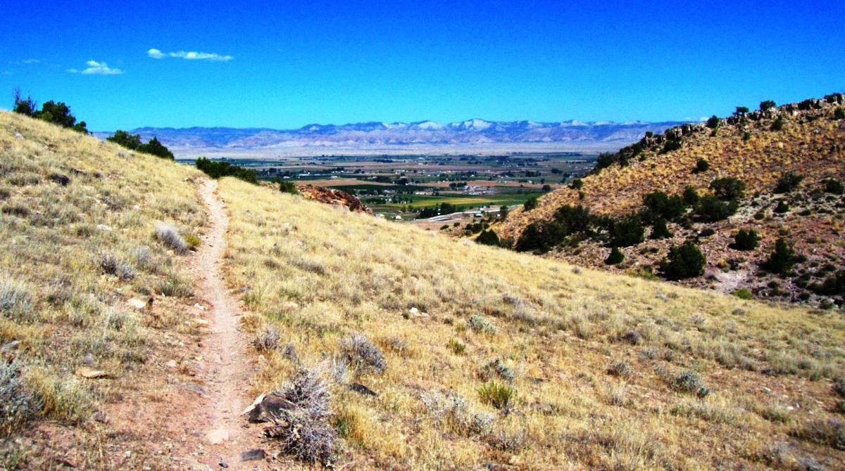 A scenic landscape featuring a winding dirt path leading downhill through golden grass and shrubbery. In the background, a lush green valley is nestled between rolling hills and majestic mountain peaks under a vibrant blue sky with a few scattered clouds. Moore Fun mountain bike trail.