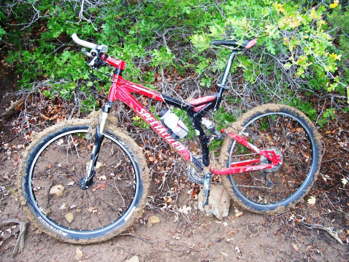 A red and black mountain bike, partially covered in mud, is resting against a bush in a forested area. The ground is sandy with fallen leaves, and the bike's wheels have accumulated dirt, suggesting recent use on rugged terrain. Telegraph Trail System mountain bike trail.