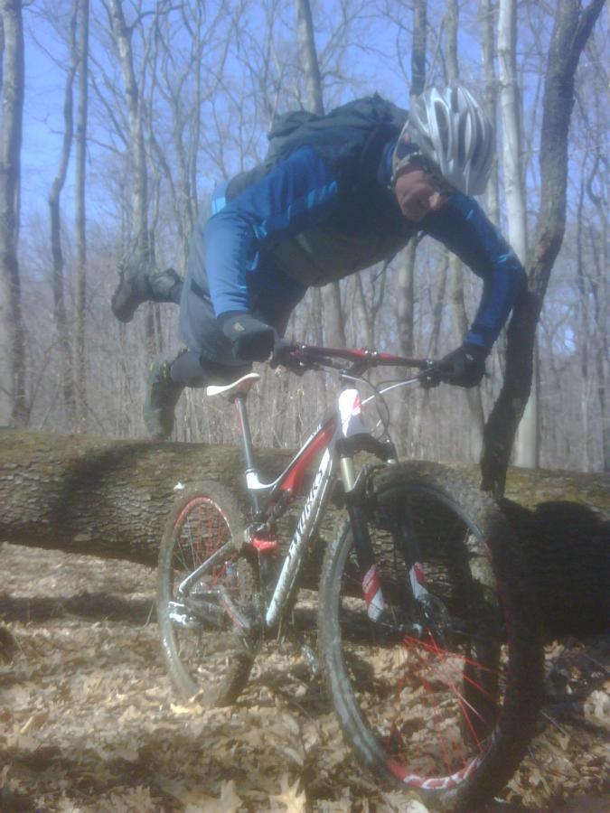 A mountain biker performing a stunt while navigating over a fallen tree in a wooded area, with trees and blue sky in the background. The rider is in an athletic pose, wearing a helmet and gear, and has a backpack on. The bike is balanced precariously on the log. Hartwood Acres mountain bike trail.