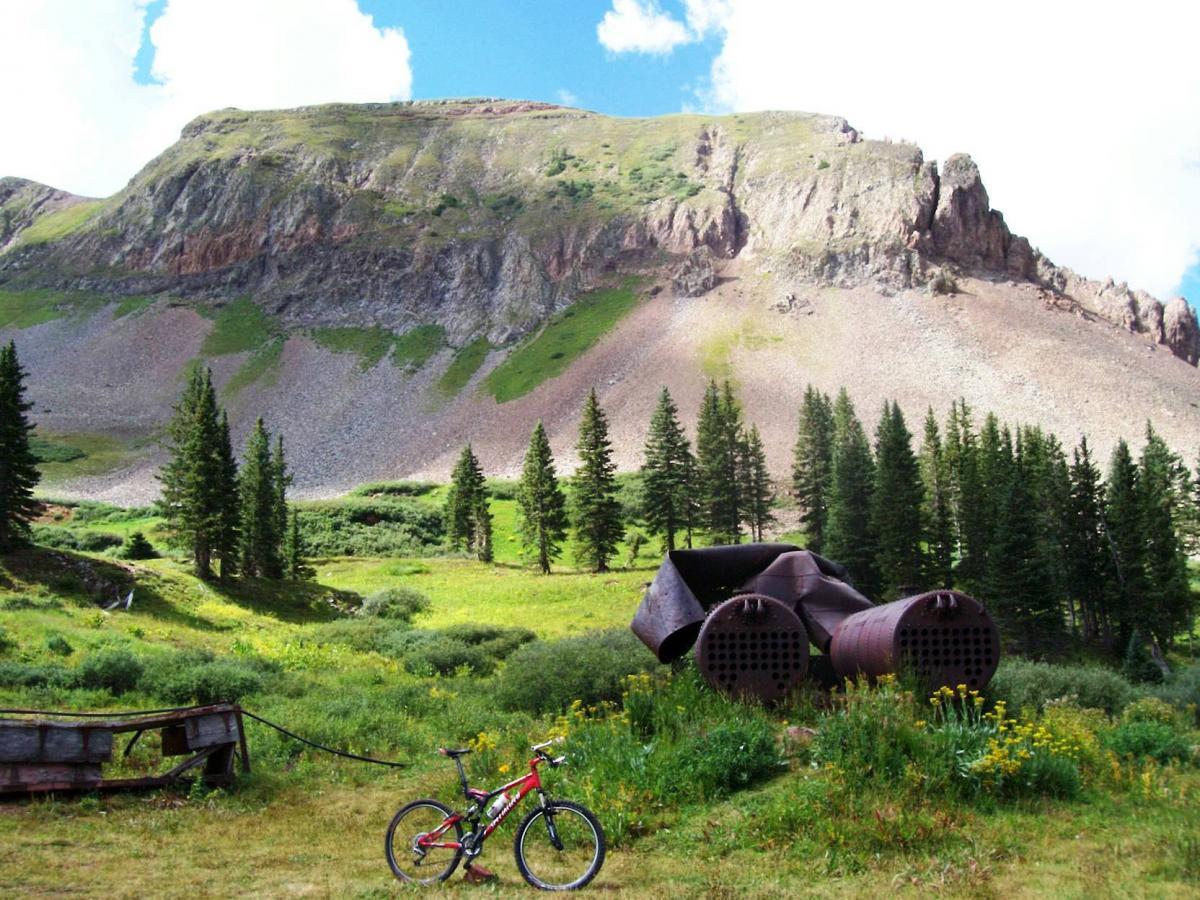 A serene landscape featuring a mountainous backdrop with lush greenery, pine trees, and wildflowers. In the foreground, a red mountain bike leans against a rusty, old industrial structure partially hidden by grass. A wooden cart is also visible nearby, set against the vibrant natural scenery. The sky above is blue with fluffy white clouds. Colorado Trail: Kennebec Pass To Junction Creek mountain bike trail.