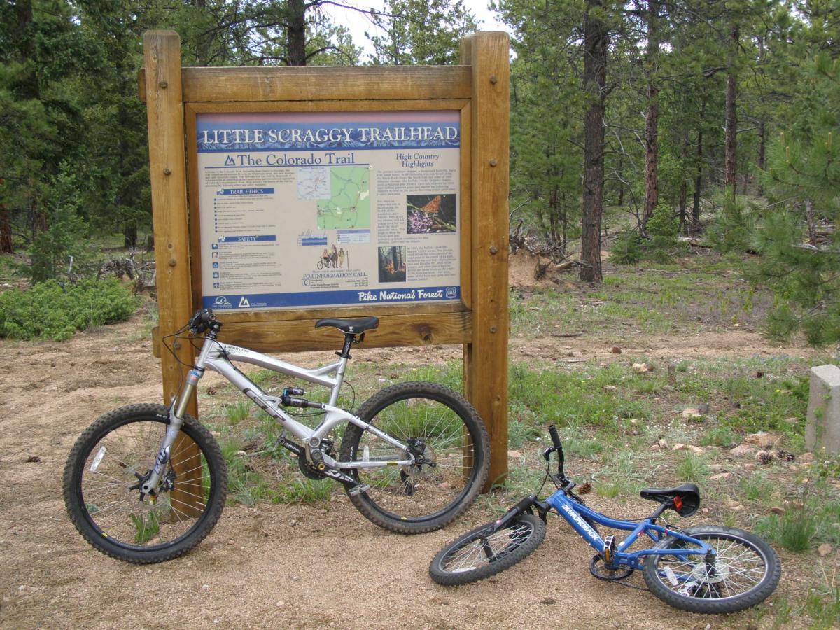A wooden trailhead sign for Little Scraggy Trail, featuring maps and information about the Colorado Trail. In the foreground, a silver mountain bike is leaning against the sign, alongside a small blue children's bike. The surrounding area is wooded, with pine trees and a dirt path. Buffalo Creek mountain bike trail.