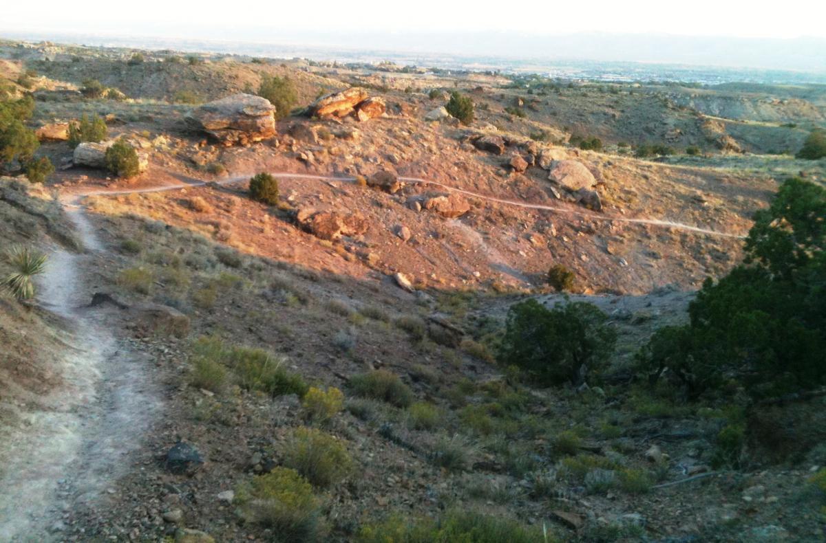 A scenic view of a rugged landscape featuring a winding dirt path leading through rocky terrain, with patches of green shrubs and small trees scattered across the hillside. The setting sun casts warm light on the rocks, highlighting their textures against the backdrop of distant mountains and a clear sky. Lunch Loops mountain bike trail.