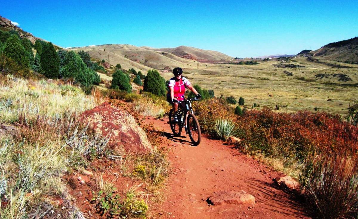 A mountain biker riding along a dirt trail surrounded by scenic hills and greenery under a clear blue sky. The trail features reddish dirt and sparse vegetation, creating a beautiful outdoor landscape suitable for biking. Red Rocks / Dakota Ridge mountain bike trail.