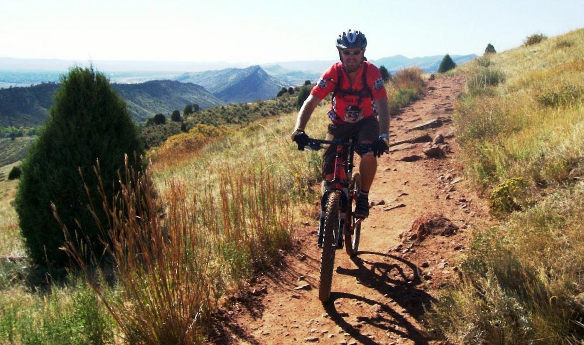 A mountain biker riding along a rocky trail surrounded by tall grass and small shrubs, with scenic hills and valleys in the background under a clear blue sky. Red Rocks / Dakota Ridge mountain bike trail.