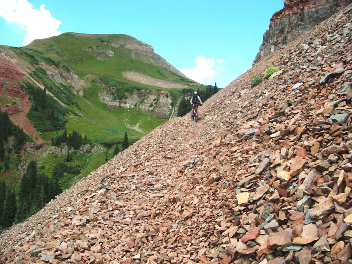 A mountain biker navigates a rocky trail on a sloped terrain surrounded by lush green hills and a clear blue sky. Colorado Trail: Kennebec Pass To Junction Creek mountain bike trail.