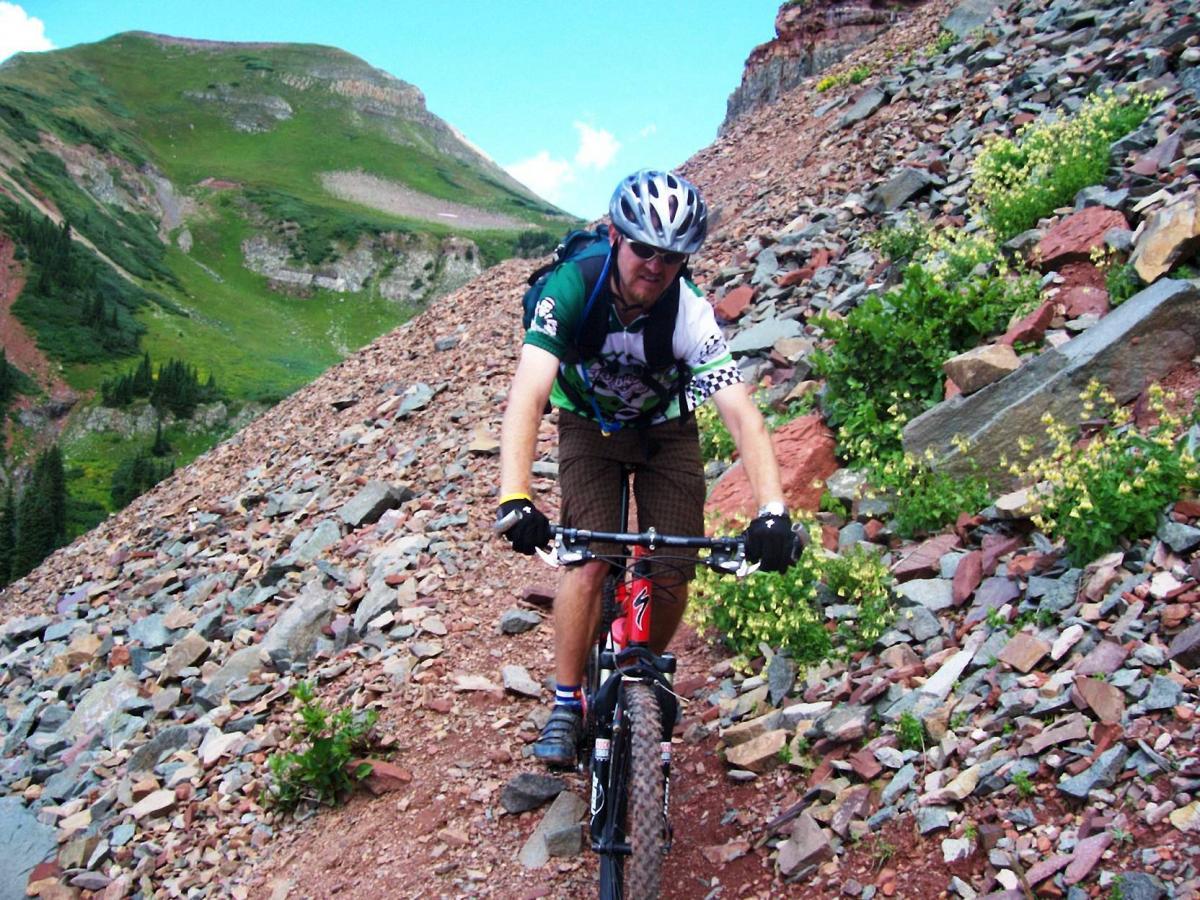A mountain biker navigating a rocky trail surrounded by green hills and sparse vegetation under a blue sky. The cyclist is wearing a helmet and protective gear, focused on maintaining balance while riding uphill. Colorado Trail: Kennebec Pass To Junction Creek mountain bike trail.